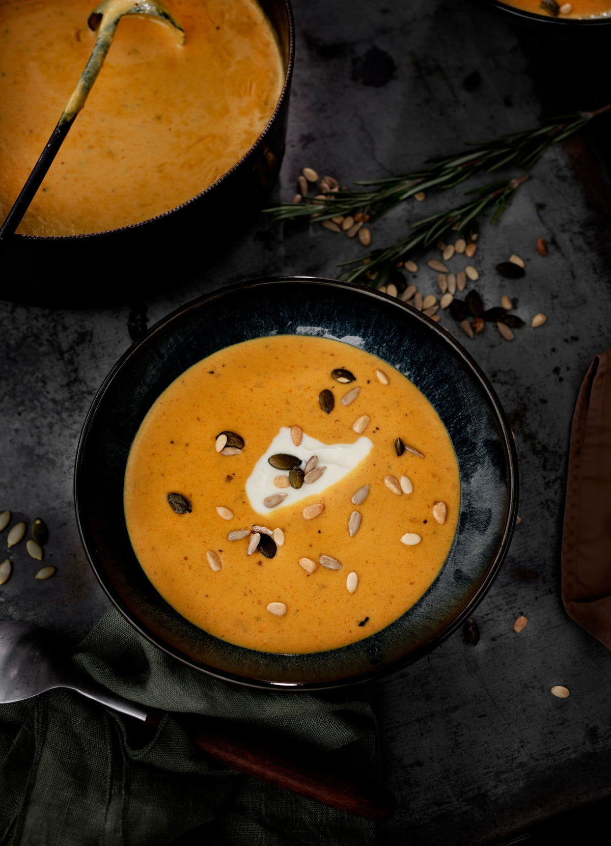 Bowl of winter pumpkin soup with crème fraîche and toasted seed mix, next to a pot of soup with a ladle